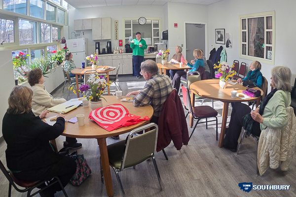 A group of people sits at round tables in a bright, sunlit room, attentively listening to a person standing at the front.